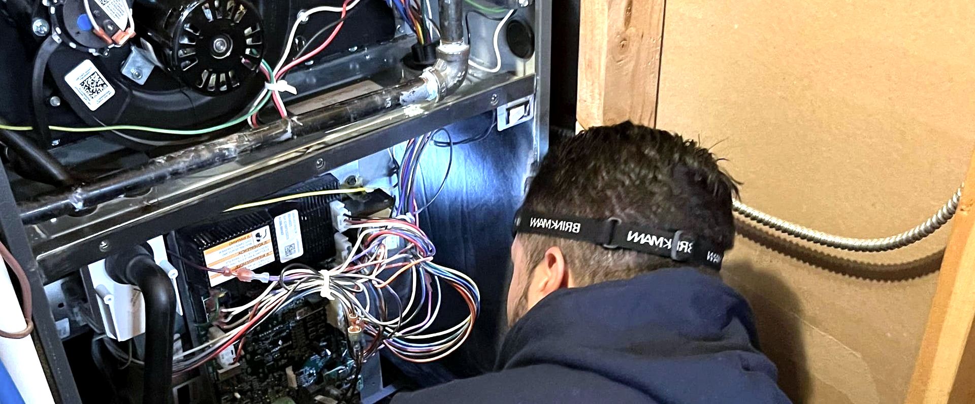 Technician crouched beside open furnace with wiring, headlamp on head
