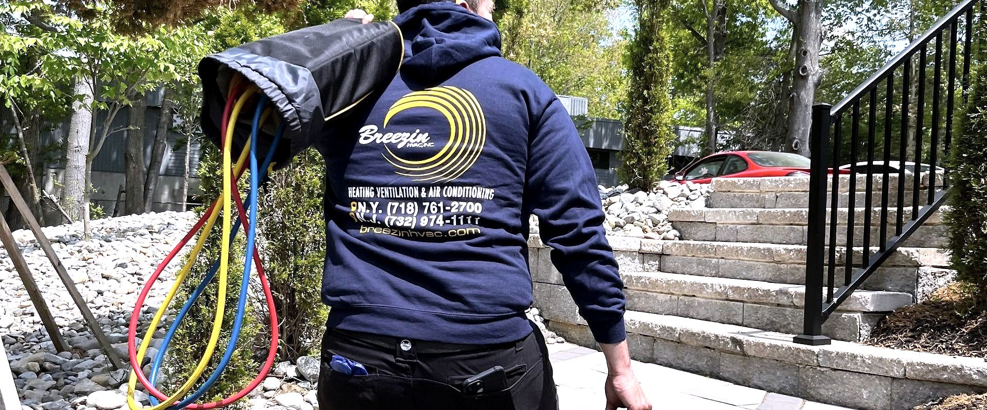 Technician in Breezin hoodie carrying coiled refrigerant lines past stone steps